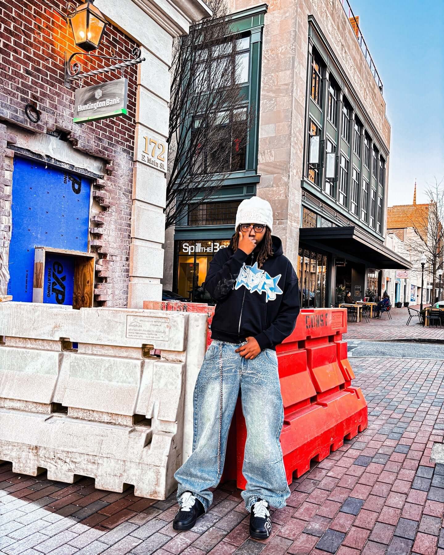 Model wearing a black graffiti zip-up hoodie with 3D embroidery, styled with baggy jeans and sneakers in an urban setting.