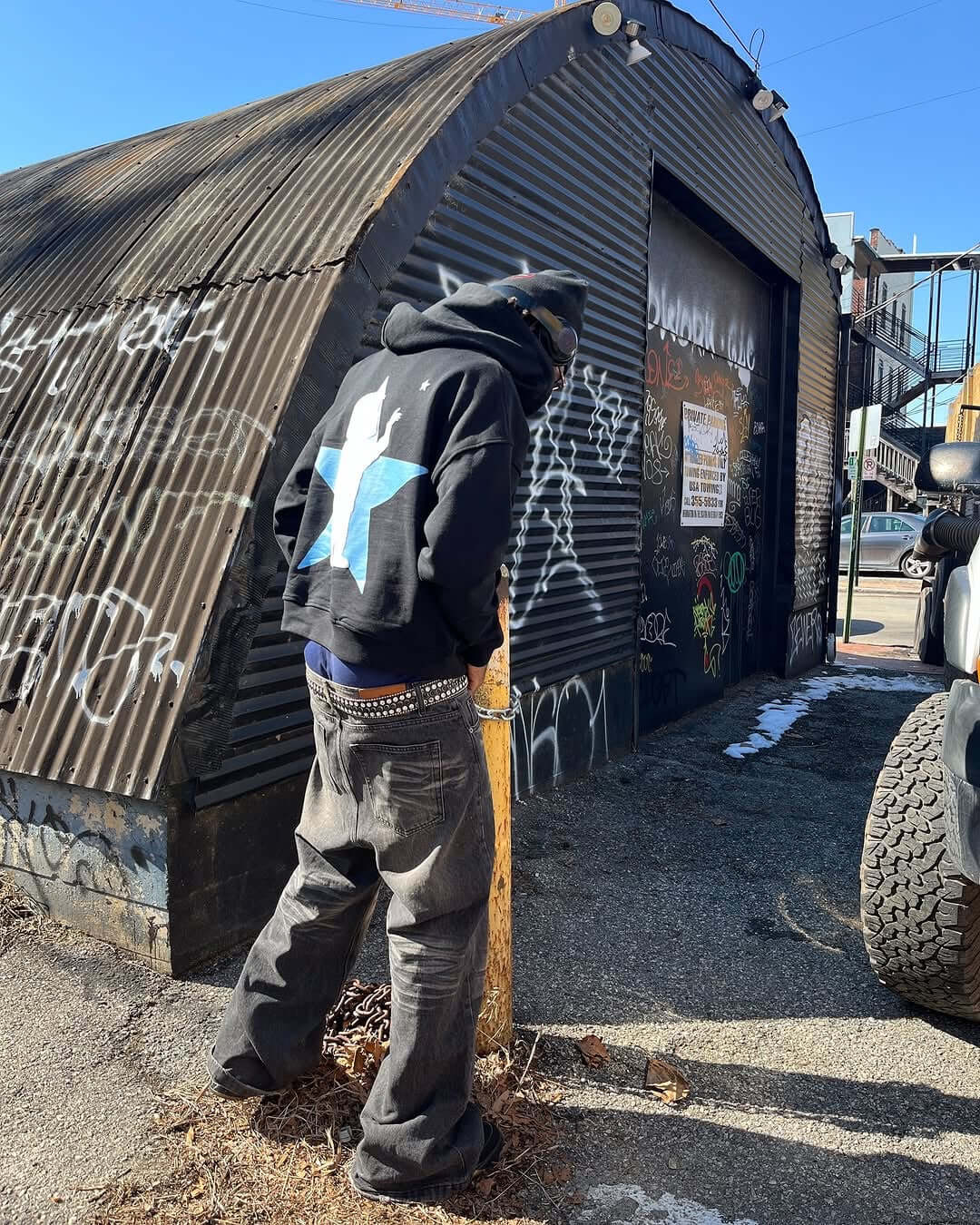 Person wearing a black graffiti zip up hoodie with a blue star logo, standing near a graffiti-covered building.