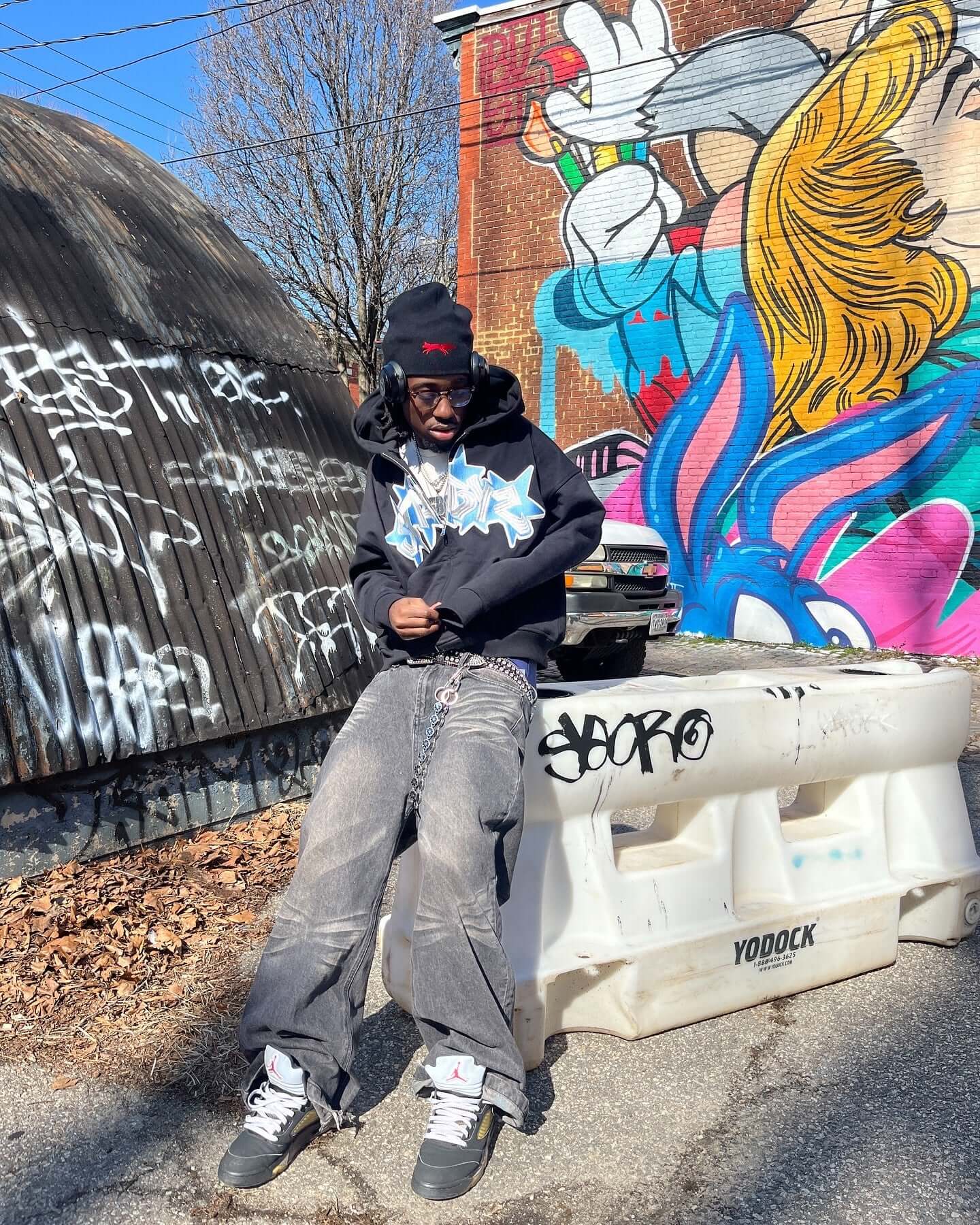 Person wearing a black graffiti zip-up hoodie in front of a colorful mural, surrounded by urban street art.