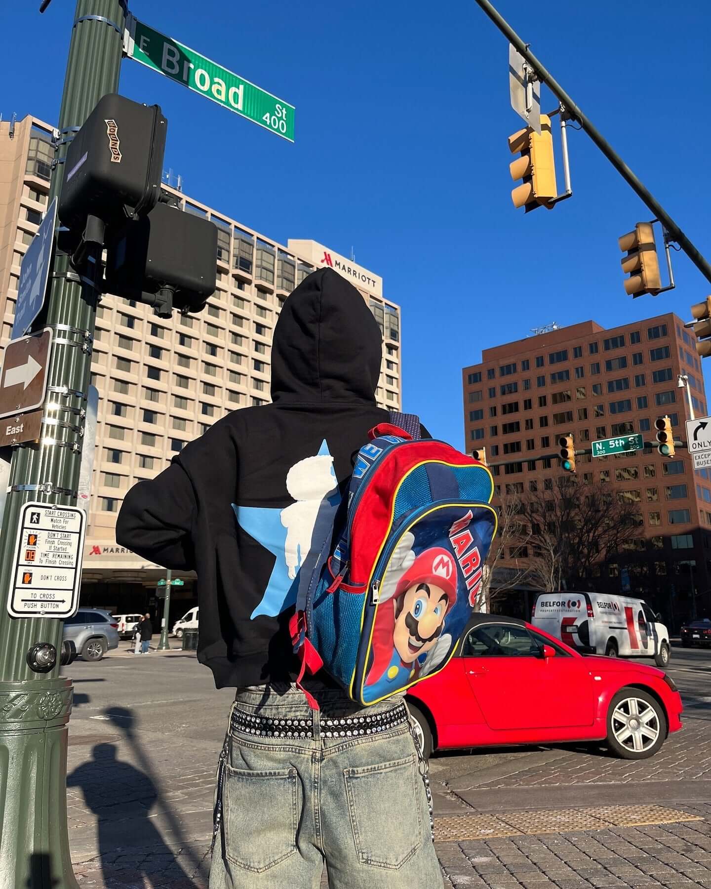 Person wearing a black hoodie with a blue star and a colorful Mario backpack at a city intersection.